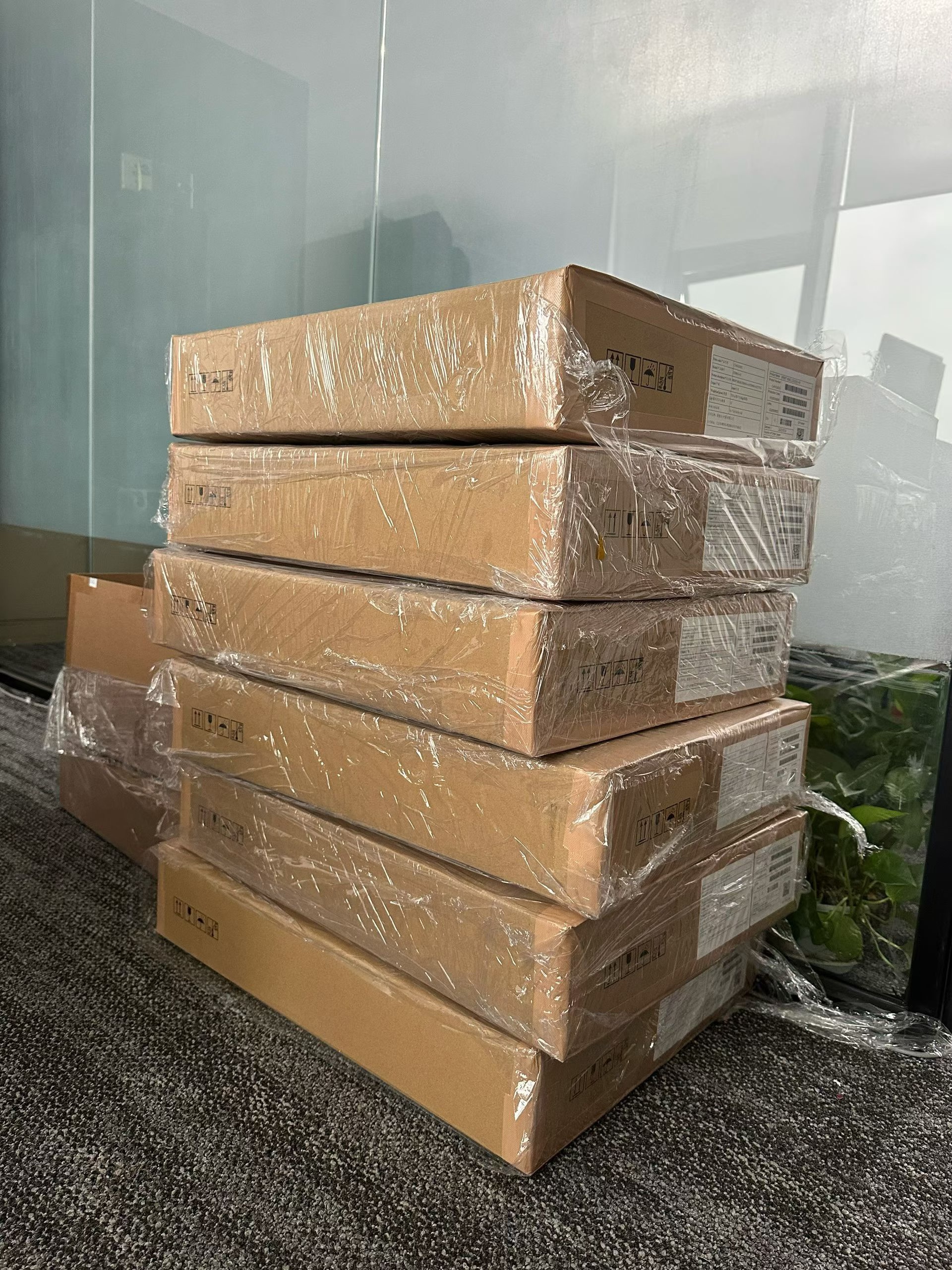 A stack of five sealed cardboard boxes wrapped in plastic, placed on a carpeted floor next to a glass wall.