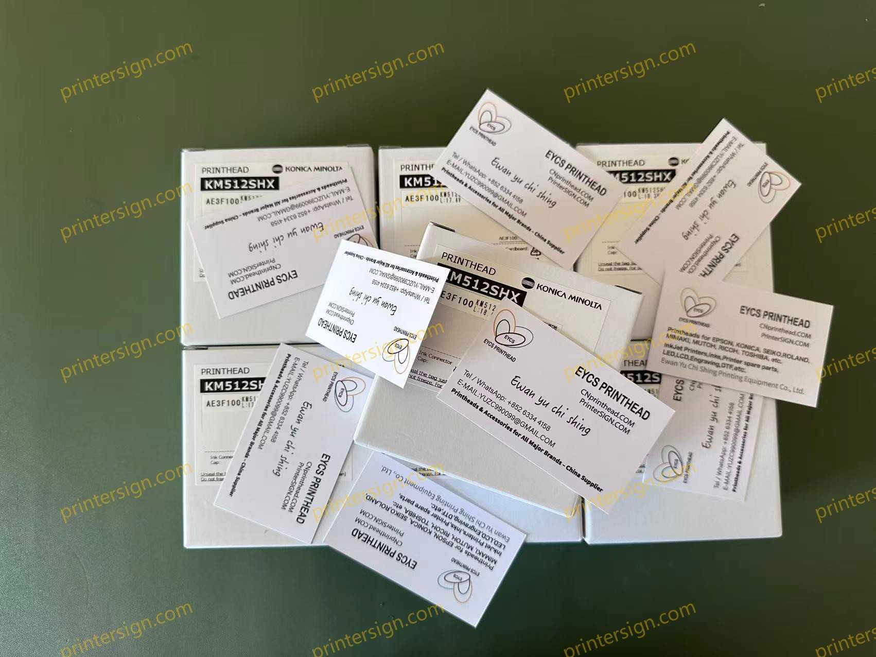 A collection of printer head boxes and business cards scattered on a surface, featuring the Konica Minolta KM512SHX model. The boxes are labeled with product information and the business cards include contact details for printing services.