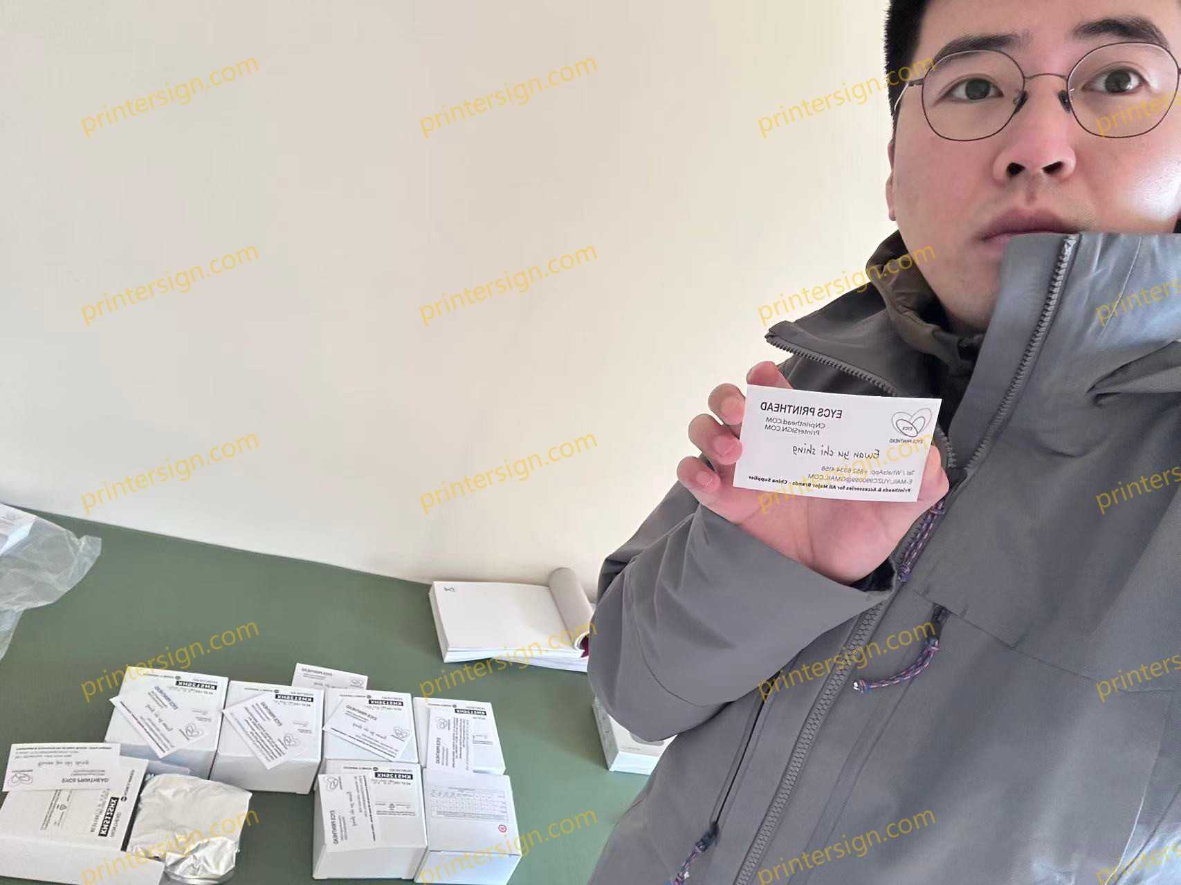 A person holding a printed card featuring a logo and text, standing in front of a table with multiple white boxes and other printing materials.