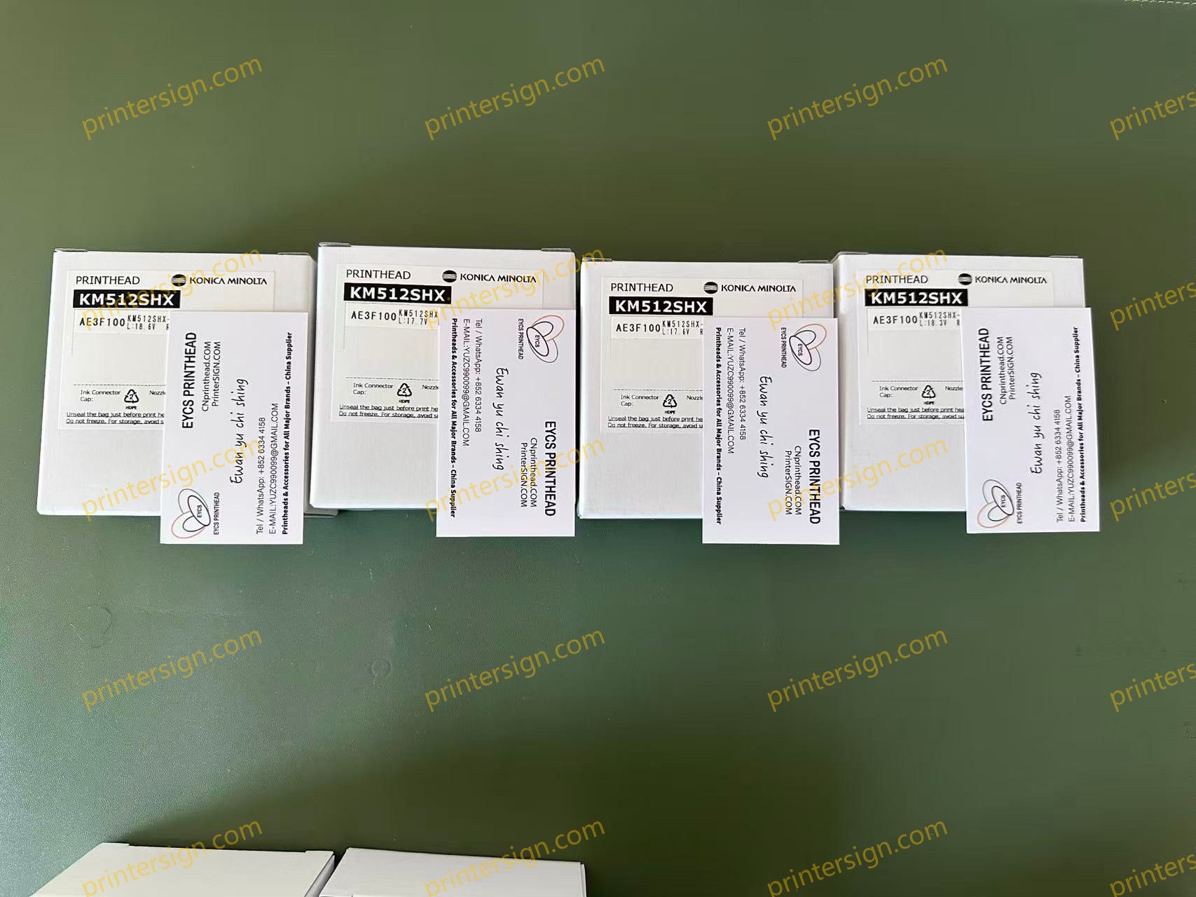 A row of four boxes labeled 'KM512SHX' containing printheads, with additional business cards placed beside them on a green surface.