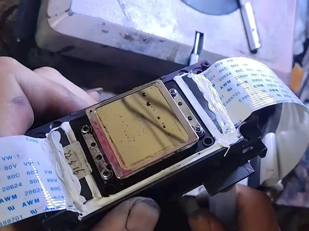 Close-up view of a hand holding a printed circuit board with a gold heat sink and flexible ribbon cable attached.